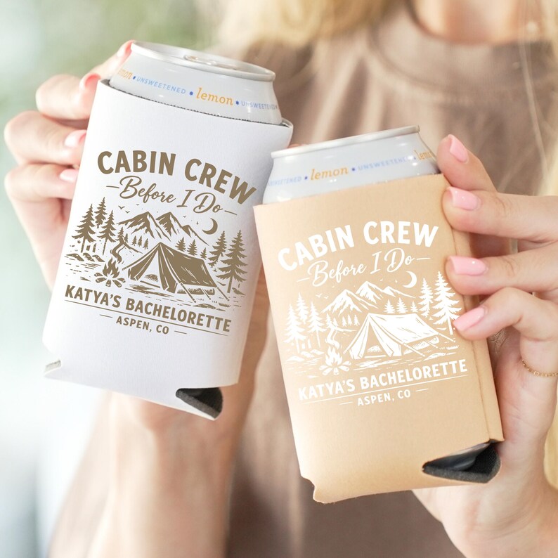 May include: Two can coolers, one white and one tan, each with the text "CABIN CREW - Before I Do - KATYA'S BACHELORETTE - ASPEN, CO" in a mountain and camping design. The coolers are holding cans.
