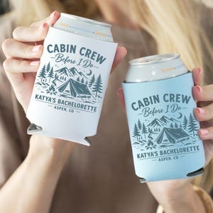 May include: Two can coolers, one white and one light blue, each with a camping-themed design. The text "CABIN CREW - Before I Do - KATYA'S BACHELORETTE - ASPEN, CO" is printed on each cooler. The design includes trees, mountains, a tent, and a campfire.