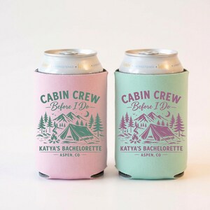 May include: Two can coolers, one pink and one teal, each featuring a camping-themed design. The text reads "CABIN CREW - Before I Do - KATYA'S BACHELORETTE - ASPEN, CO" in a forest green and purple color scheme. The design includes a tent, mountains, trees, and a campfire.