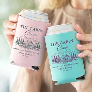 May include: Two can coolers, one pink and one teal, each with the text "THE CABIN Crew" and a cabin graphic. The text "DANIELLA'S BACHELORETTE - Aspen, Colorado - Est. 2026" is also printed on the coolers.