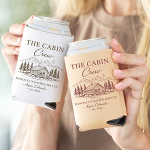 May include: Two can coolers, one white and one tan, each with the text "THE CABIN Crew" above a cabin illustration. The text "DANIELLA'S BACHELORETTE - Aspen, Colorado - Est. 2026" is printed below the cabin design.