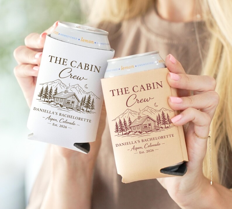 May include: Two can coolers, one white and one tan, each with the text "THE CABIN Crew" above a cabin illustration. The text "DANIELLA'S BACHELORETTE - Aspen, Colorado - Est. 2026" is printed below the cabin design.