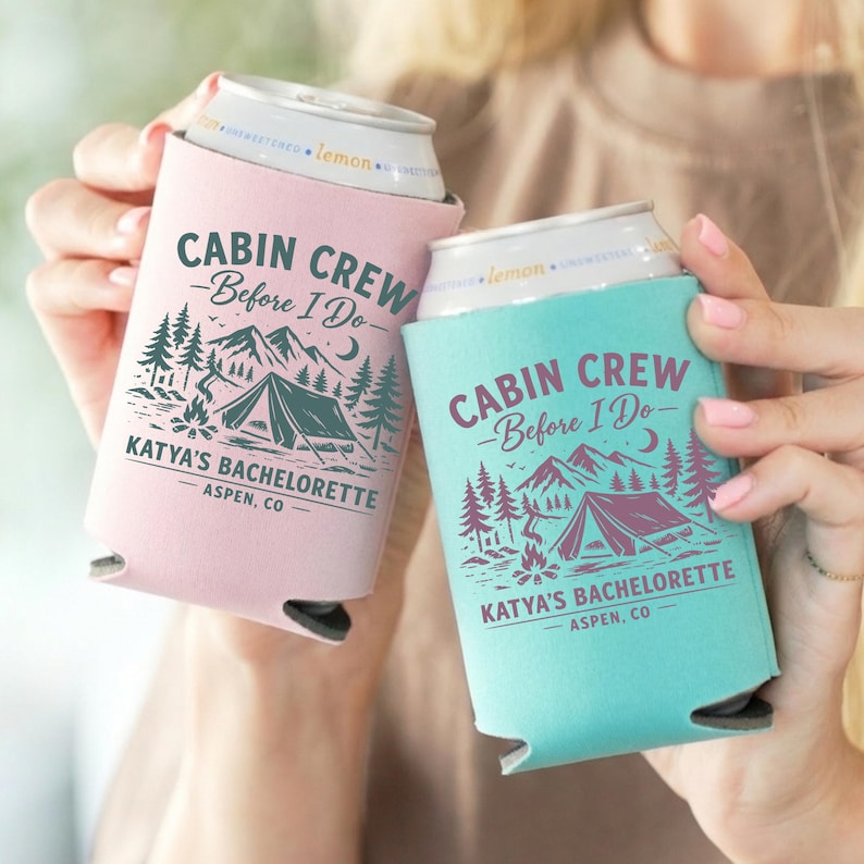 May include: Two can coolers, one pink and one teal, each with a camping-themed design. The text on the coolers reads "CABIN CREW - Before I Do - KATYA'S BACHELORETTE - ASPEN, CO". The design includes mountains, trees, a tent, and a campfire.
