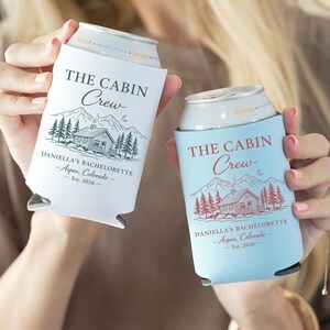 May include: Two can coolers, one white and one light blue, each with the text "THE CABIN Crew" and "DANIELLA'S BACHELORETTE - Aspen, Colorado - Est. 2026". The design includes a cabin, mountains, trees, and a crescent moon.