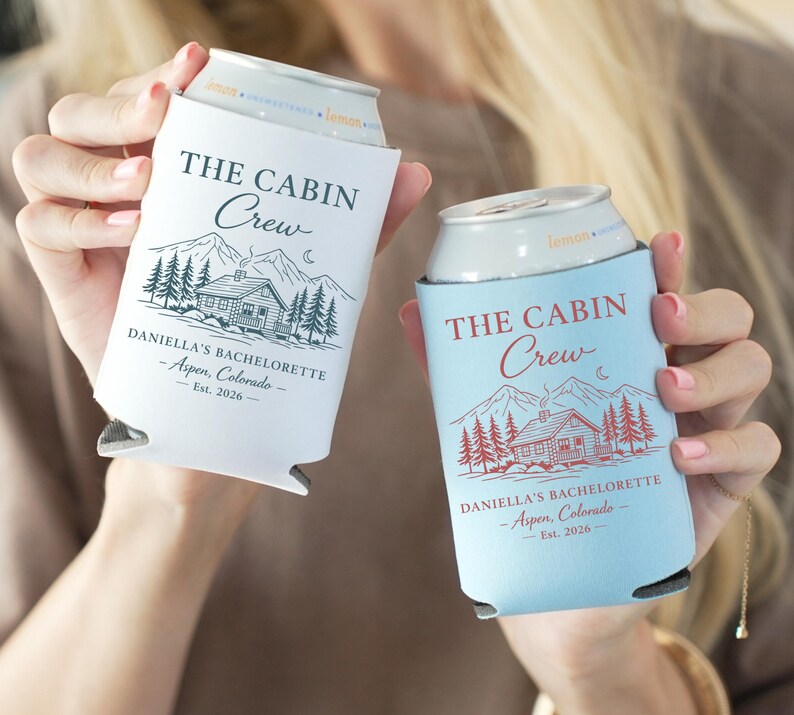 May include: Two can coolers, one white and one light blue, each with the text "THE CABIN Crew" and "DANIELLA'S BACHELORETTE - Aspen, Colorado - Est. 2026". The design includes a cabin, mountains, trees, and a crescent moon.