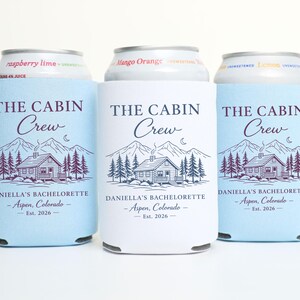 May include: Three drinks can coolers with the text "THE CABIN Crew" and a cabin graphic. The coolers are light blue and white, with the text "DANIELLA'S BACHELORETTE - Aspen, Colorado - Est. 2026".
