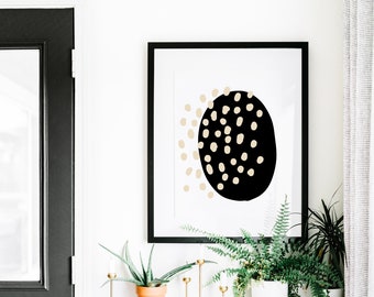 Puede incluir: Una impresión abstracta en blanco y negro con un gran círculo negro con lunares beige. La impresión está enmarcada en un marco negro y está colgada en una pared blanca sobre un armario blanco con dos cajones. Hay dos plantas en maceta en el armario, una con hojas verdes y otra con una suculenta verde.