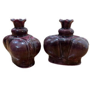 May include: Two antique, dark purple glass perfume bottles. Each bottle has a bulbous body with a decorative pattern and a crown-shaped stopper. The bottles are displayed against a white background.