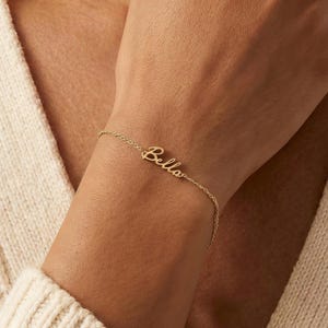 Może przedstawiać: Delikatna, złota bransoletka z napisem "Bella" w ozdobnej czcionce. Delikatny łańcuszek podkreśla imię. Biżuteria jest noszona na nadgarstku, co sugeruje spersonalizowany dodatek.