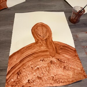 Puede incluir: Pintura de acuarela marrón sobre papel que representa una figura con la cabeza con capucha y cabello suelto. La obra de arte tiene una apariencia texturizada y en capas. Se ven otras pinturas y un vaso de pintura.