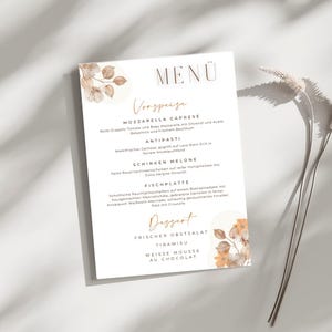 Könnte beinhalten: Eine gedruckte Menükarte mit einem floralen Design in Braun- und Goldtönen. Die Speisekarte enthält Gerichte wie Mozzarella Caprese und Tiramisu, mit Text auf Deutsch. Die Karte ist rechteckig.