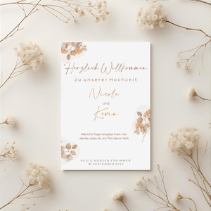 Könnte beinhalten: Eine rechteckige Hochzeitseinladung mit dem Text "Herzlich Willkommen" und den Namen Nicole und Kevin in goldener Schrift. Die Einladung ist von getrockneten Blumen und Zweigen umgeben, mit dem Datum 20. September 2025.