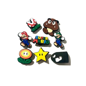 May include: A collection of eight colourful rubber shoe charms featuring Super Mario characters and icons, including Mario, Luigi, a Piranha Plant, a Goomba, a Fire Flower, a yellow star, a Bullet Bill, and a Super Mario logo in blue, red, and yellow.