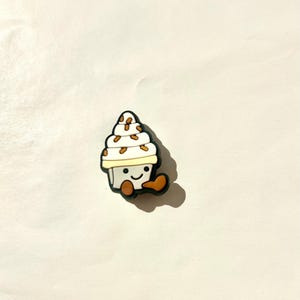 Pu&ograve; includere: Un ciondolo a forma di cono gelato bianco e beige con un volto sorridente e dettagli marroni. Il ciondolo ha un contorno nero e si trova su uno sfondo bianco. Il cono gelato ha delle gocce marroni.