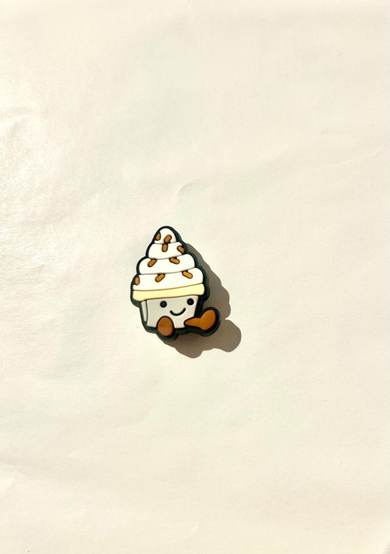 Pu&ograve; includere: Un ciondolo a forma di cono gelato bianco e beige con un volto sorridente e dettagli marroni. Il ciondolo ha un contorno nero e si trova su uno sfondo bianco. Il cono gelato ha delle gocce marroni.