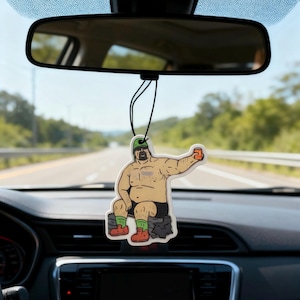 Peut inclure: Un désodorisant de voiture fantaisie représentant un personnage de dessin animé tenant une chope de bière. Le désodorisant est suspendu au rétroviseur d'une voiture. Le personnage est bronzé avec un short noir, un chapeau vert, des bottes rouges et des chaussettes vertes.