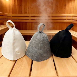 Könnte beinhalten: Drei Saunahüte in Weiß, Grau und Schwarz, jeweils mit einer Schlaufengriff. Die Hüte werden auf einer Holzbank in einer Sauna ausgestellt, mit Dampf im Hintergrund. Die Hüte sollen den Kopf vor Hitze schützen.