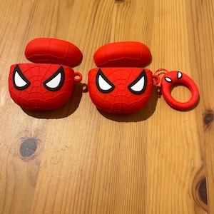 May include: Two red Spider-Man-themed AirPod cases with matching lids and a small, round keychain. The cases feature a web design and white eye details. The items are displayed on a wooden surface.