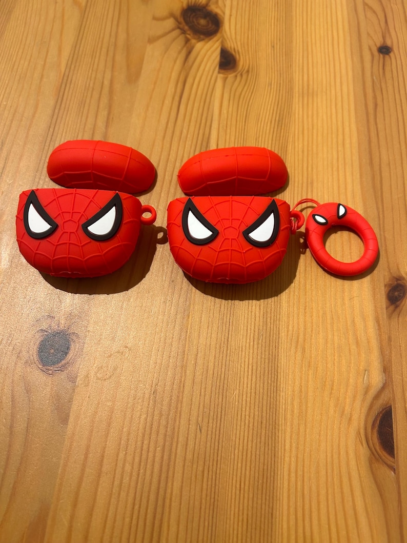 May include: Two red Spider-Man-themed AirPod cases with matching lids and a small, round keychain. The cases feature a web design and white eye details. The items are displayed on a wooden surface.