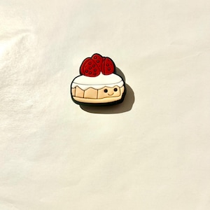 Pu&ograve; includere: Un ciondolo per scarpe colorato a forma di cheesecake a cartoni animati con una faccia sorridente. La cheesecake ha una parte superiore bianca, una base marrone ed &egrave; sormontata da tre fragole rosse.