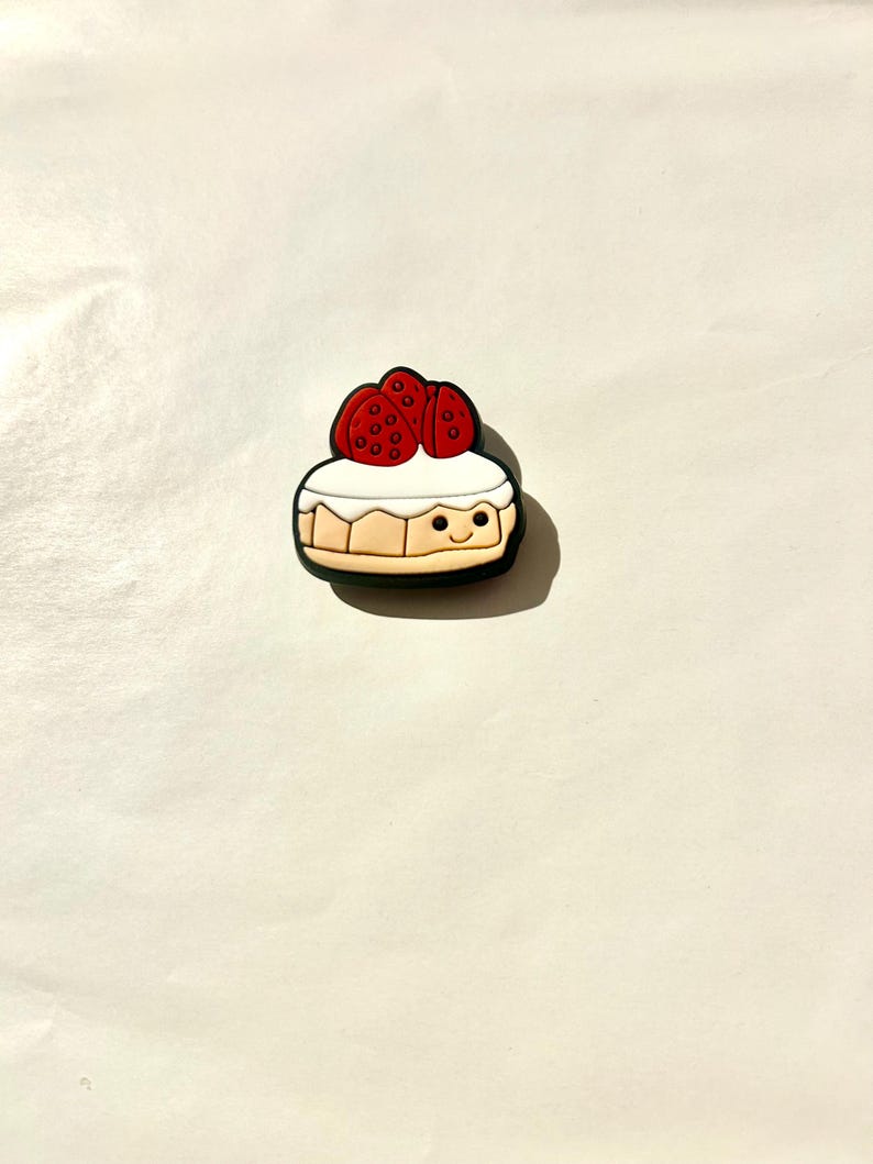 Pu&ograve; includere: Un ciondolo per scarpe colorato a forma di cheesecake a cartoni animati con una faccia sorridente. La cheesecake ha una parte superiore bianca, una base marrone ed &egrave; sormontata da tre fragole rosse.