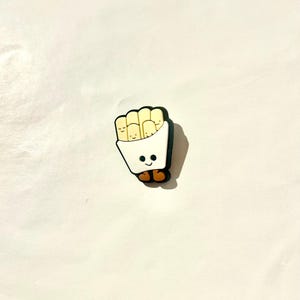 Pu&ograve; includere: Un ciondolo per scarpe a forma di contenitore di patatine fritte sorridente. Il contenitore &egrave; bianco con un contorno nero e contiene diverse patatine fritte gialle con facce sorridenti. Il ciondolo ha piedi marroni e una base nera.