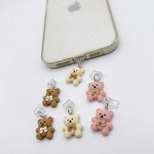 Puede incluir: Una colección de bonitos colgantes para teléfono con forma de osito de peluche en varios colores: marrón, crema y rosa. Cada colgante tiene un pequeño lazo y está unido a un enchufe de plástico transparente. También hay una funda de teléfono blanca brillante.