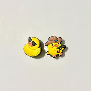 May include: Two yellow duck-shaped shoe charms. One duck wears black sunglasses and faces left. The other duck wears a brown cowboy hat with a blue band and holds a black handgun. Both charms feature vibrant yellow bodies with orange beaks and feet.