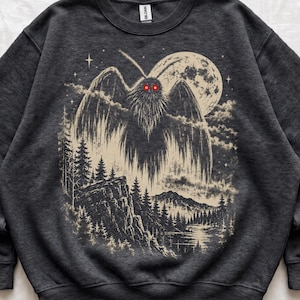 May include: A dark heather gray crewneck sweatshirt featuring a graphic print of a moth-like creature with glowing red eyes hovering over a forest landscape with a large full moon, stars, and a river under a night sky.
