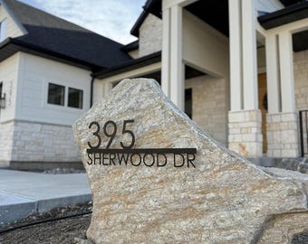 May include: A large, light-colored natural stone boulder featuring a black metal address sign. The sign displays the number 395 above the text SHERWOOD DR, separated by a horizontal line, mounted directly onto the front face of the rock.