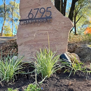 May include: A large, light brown, natural stone boulder featuring a black metal address sign mounted on its front. The sign displays the number 6795 above the word CHAPARRAL in a clean, sans-serif font, creating a clear house marker.