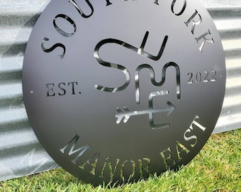 May include: Black metal sign with the text "South Fork Manor East" and a stylized "SM" logo with an arrow pointing to the right. The sign also includes the text "Est. 2022".