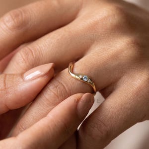May include: A gold ring featuring a gentle wave-shaped band with a central round light blue gemstone flanked by two smaller clear stones. The metal has a polished finish, and the design follows a smooth, curved contour along the finger.