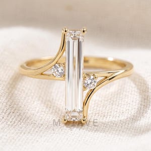 May include: A gold ring featuring a central, vertical, rectangular clear gemstone. Two smaller, round, clear gemstones are set either side of the central stone. The ring is set on white fabric.