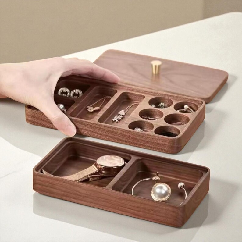 K&ouml;nnte beinhalten: Eine Holzschmuckschatulle mit Scharnierdeckel und mehreren F&auml;chern. Die Box ist ge&ouml;ffnet und zeigt verschiedene Schmuckst&uuml;cke, darunter Ohrringe, Halsketten und Ringe. Eine goldfarbene Uhr und ein Armband mit einer Perle sind ebenfalls sichtbar.