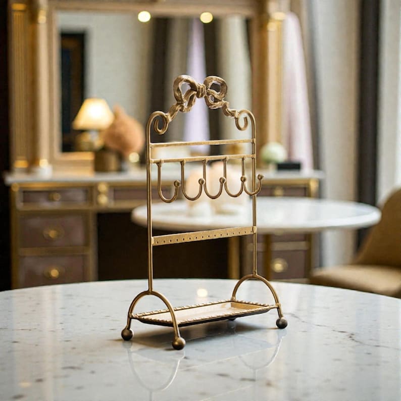 May include: A gold-colored metal jewelry organizer with a bow-shaped top. The organizer has hooks for necklaces and a shelf for small items. It stands on a marble table, showcasing a vintage aesthetic.