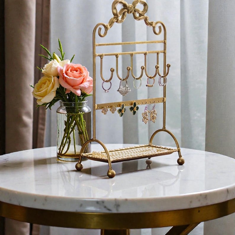 May include: A gold-colored jewelry organizer with a bow-shaped top. The organizer has hooks for rings and earrings, and a small shelf. A vase of flowers sits to the left of the organizer on a marble table.