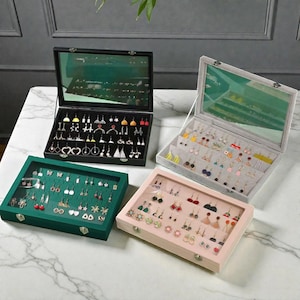 May include: Four earring display cases in black, green, pink, and gray, each filled with various earrings. The cases have glass lids and are arranged on a white marble surface. The earrings are in different styles and colors.
