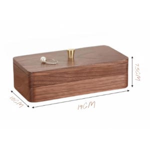 K&ouml;nnte beinhalten: Eine rechteckige Schmuckschatulle aus Holz mit dunkelbraunem Finish. Die Box ist 7,5 cm hoch, 10 cm breit und 19 cm lang. Sie hat einen goldfarbenen Knopf oben und einen Ring mit einer Perle auf dem Deckel.