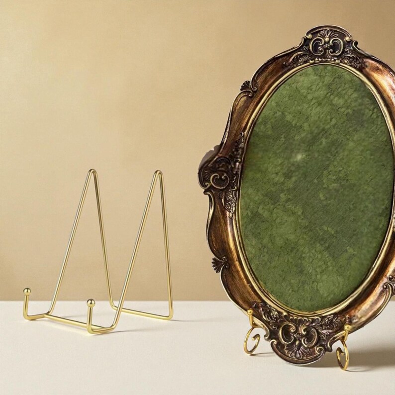 May include: An ornate, oval-shaped picture frame with a dark brown, gold-trimmed border and a green interior. A gold-colored metal display stand is next to the frame. The frame is supported by a small gold stand.