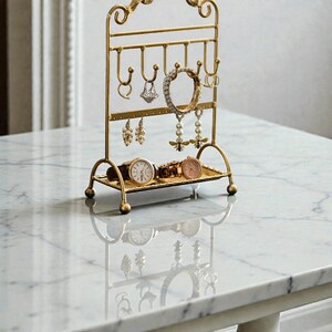May include: A gold-colored metal jewelry organizer with a decorative top and curved base. The organizer holds various earrings, rings, bracelets, and watches. The watches are displayed on a small tray at the base of the organizer.