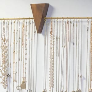 May include: A wooden jewellery organiser with a brass-coloured bar and numerous necklaces hanging from it. The organiser has a triangular wooden piece at the top, and the necklaces are of various styles and lengths.