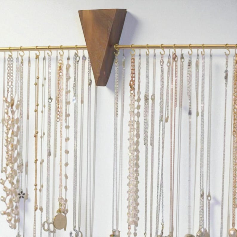 May include: A wooden jewellery organiser with a brass-coloured bar and numerous necklaces hanging from it. The organiser has a triangular wooden piece at the top, and the necklaces are of various styles and lengths.