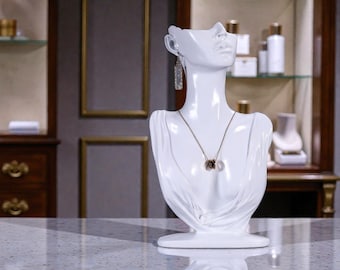 May include: A white jewellery display bust, featuring a necklace with three circular pendants and dangling earrings. The bust is on a reflective surface, with a blurred background of a shop interior. The bust is designed to display necklaces and earrings.