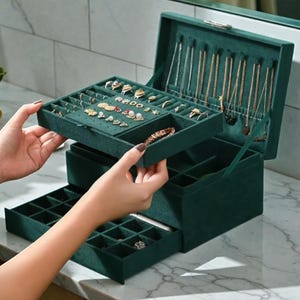 Può includere: Un portagioie in velluto verde scuro con più scomparti e cassetti. La scatola è aperta, rivelando anelli, collane e braccialetti. La scatola è progettata per riporre e organizzare i gioielli. La scatola è su una superficie di marmo.