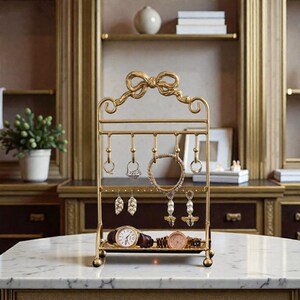 May include: A gold-colored jewelry organizer with a bow-shaped top. The organizer has multiple hooks for hanging necklaces, earrings, and rings. Two watches are displayed on the bottom tray. The background includes a marble surface and a wooden cabinet.