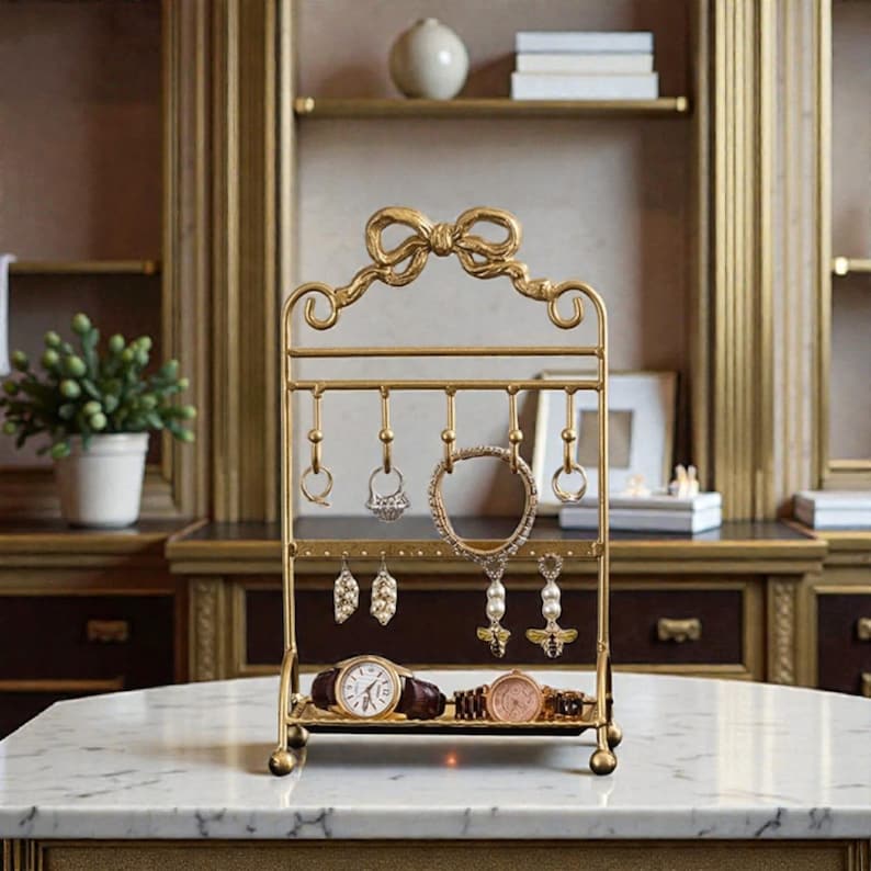 May include: A gold-colored jewelry organizer with a bow-shaped top. The organizer has multiple hooks for hanging necklaces, earrings, and rings. Two watches are displayed on the bottom tray. The background includes a marble surface and a wooden cabinet.