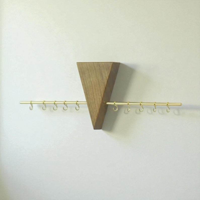 May include: A wooden wall-mounted jewellery organiser with a triangular design. The organiser features a natural wood finish and a horizontal brass bar with small hooks for hanging necklaces or bracelets. The design is minimalist and modern.
