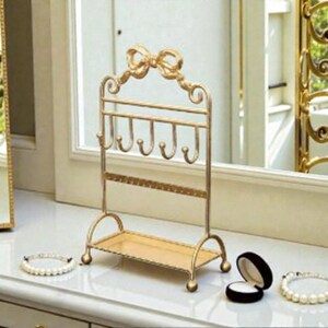 May include: A gold-colored jewelry organizer with a bow-shaped top and multiple hooks for necklaces and bracelets. The organizer has a small shelf at the bottom and is designed to hold earrings. Two pearl bracelets and a ring box are also visible.