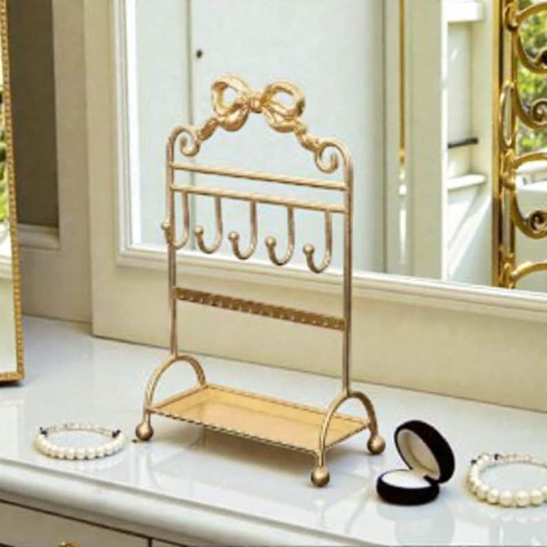 May include: A gold-colored jewelry organizer with a bow-shaped top and multiple hooks for necklaces and bracelets. The organizer has a small shelf at the bottom and is designed to hold earrings. Two pearl bracelets and a ring box are also visible.